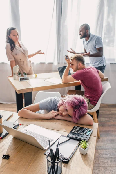 Selective focus of sad businesswoman sitting at table with laptop and stationery while multicultural colleagues working at background — Stock Photo