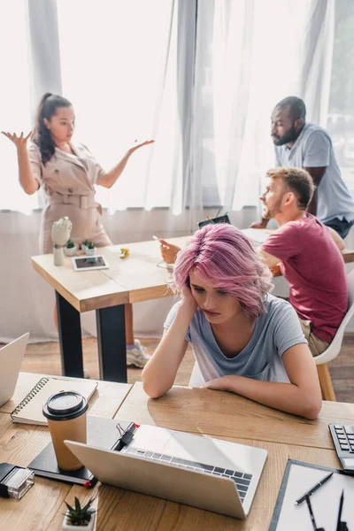 Селективный фокус расстроенной деловой женщины, сидящей за столом, в то время как коллеги-мультикультурники разговаривают в офисе — стоковое фото