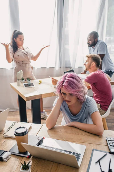 Selektiver Fokus verärgerter Geschäftsfrau, die am Tisch sitzt, während multikulturelle Mitarbeiter im Büro reden — Stockfoto