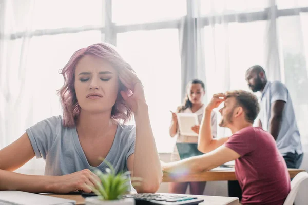 Селективный фокус разочарованной деловой женщины, сидящей за столом, в то время как коллеги-мультикультурники работают в офисе — стоковое фото