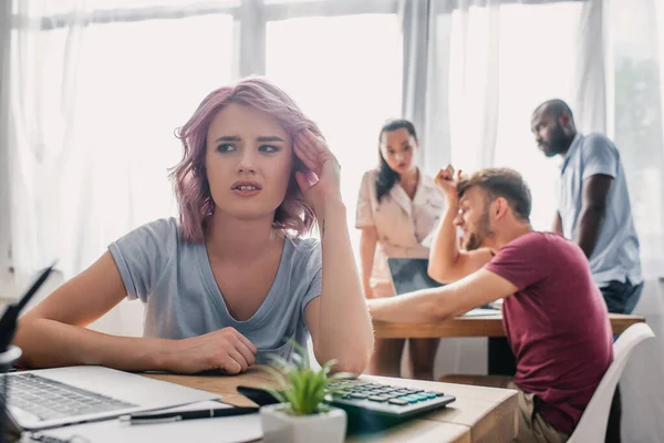 Selektiver Fokus beleidigter Geschäftsfrau, die am Tisch sitzt, während multiethnische Kollegen im Hintergrund im Büro reden — Stockfoto