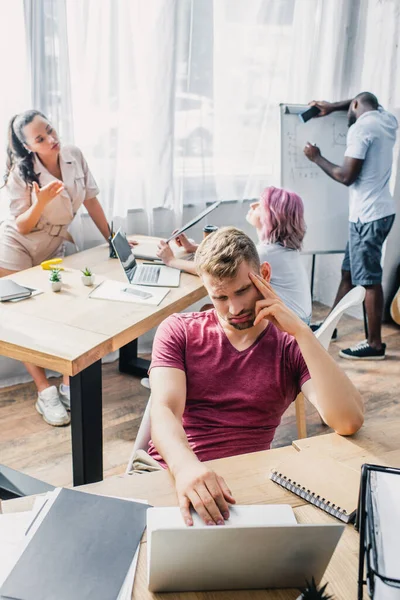 Selective focus of tired businessman using laptop near multiethnic colleagues suffering from heat in office — Stock Photo