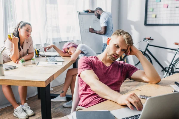 Selective focus of young businessman working on laptop while multicultural coworkers feeling hot in office — Stock Photo