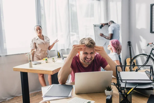 Selective focus of tired businessman looking at laptop while multiethnic colleagues suffering from heat in office — Stock Photo