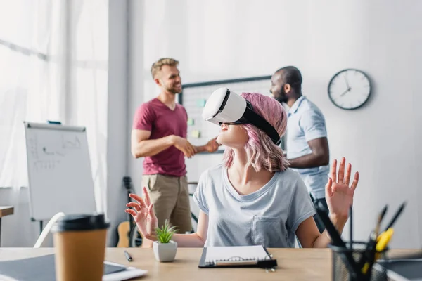 Selektiver Fokus der Geschäftsfrau mit vr-Headset am Tisch, während multiethnische Geschäftsleute im Hintergrund im Büro reden — Stockfoto