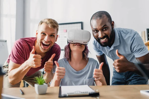 Selective focus of multiethnic business people with vr headset showing thumbs up at camera in office — Stock Photo