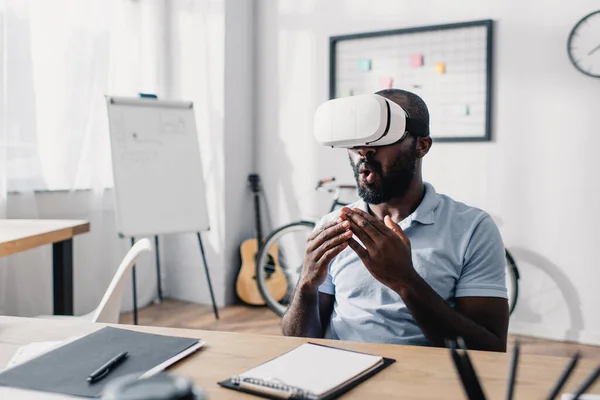 Foco seletivo do empresário afro-americano animado usando fones de ouvido vr à mesa no escritório — Fotografia de Stock