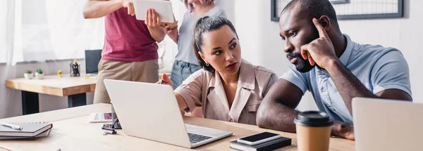 Panoramic shot of businesswoman looking at pensive african american colleague near gadgets on table in office — Stock Photo
