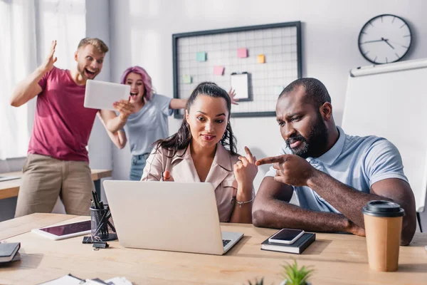 Enfoque selectivo de las personas de negocios multiculturales que trabajan con el ordenador portátil mientras sus colegas tienen videollamada en la oficina - foto de stock