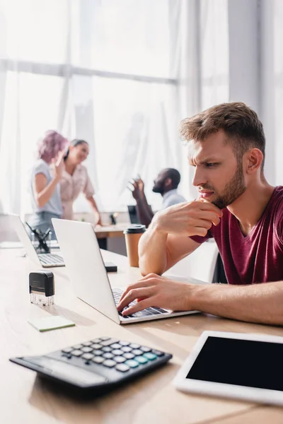 Focus selettivo dell'uomo d'affari pensoso che utilizza il computer portatile mentre i colleghi parlano in ufficio — Foto stock