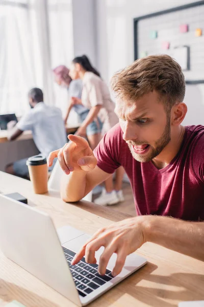 Selektiver Fokus des fokussierten Geschäftsmannes, der mit dem Finger zeigt, während er Laptop mit Kollegen im Hintergrund benutzt — Stockfoto