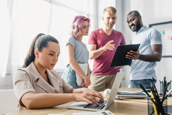 Selektiver Fokus der Geschäftsfrau mit Laptop, während multikulturelle Kollegen im Büro mit Klemmbrett arbeiten — Stockfoto