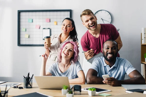 Focus selettivo di uomini d'affari multietnici con gadget e caffè per andare a guardare la fotocamera in ufficio — Foto stock