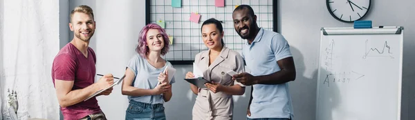 Panoramaaufnahme multikultureller Geschäftsleute mit Klemmbrettern und Gadgets, die in die Kamera im Büro blicken — Stockfoto