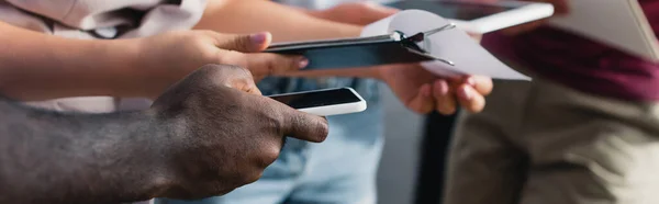 Panoramic crop of multiethnic business people holding smartphone and clipboard in office — Stock Photo