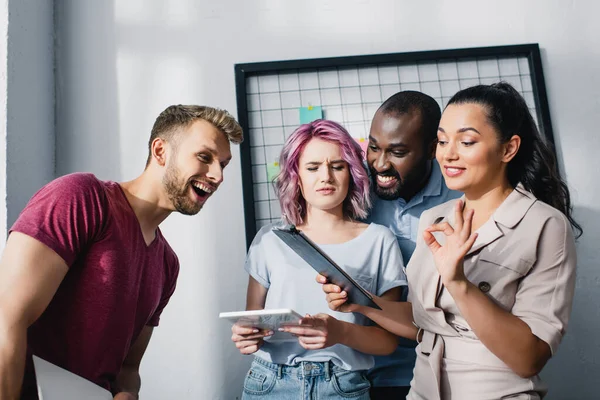 Geschäftsfrau zeigt Okay-Geste, während sie Klemmbrett in der Nähe von Kollegen im Büro hält — Stockfoto