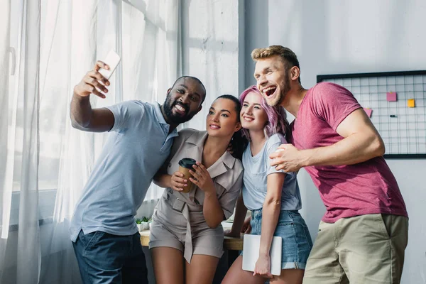 Jovens empresários multiétnicos com café para ir e gadgets tomando selfie no escritório — Fotografia de Stock