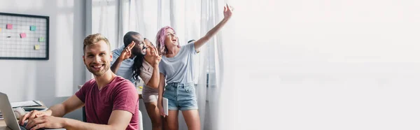 Récolte panoramique d'hommes d'affaires utilisant un ordinateur portable tout en collègues multiculturels prenant selfie dans le bureau — Photo de stock