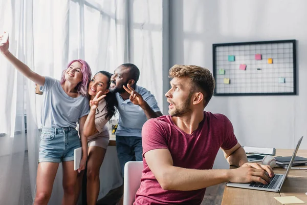 Enfoque selectivo del hombre de negocios preocupado utilizando el ordenador portátil y mirando a los colegas multiculturales tomando selfie en la oficina - foto de stock