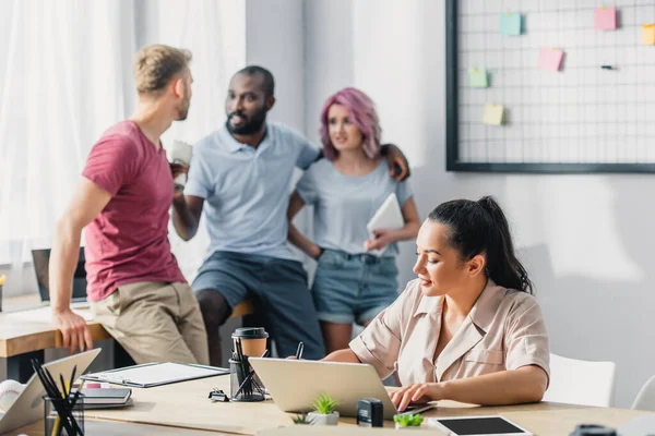 Enfoque selectivo de la mujer de negocios que usa el portátil mientras trabaja cerca de compañeros de trabajo multiétnicos en la oficina - foto de stock