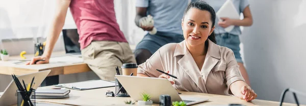 Panoramaaufnahme einer Geschäftsfrau, die in die Kamera blickt, während sie ihren Stift in der Nähe des Laptops auf dem Tisch im Büro hält — Stockfoto