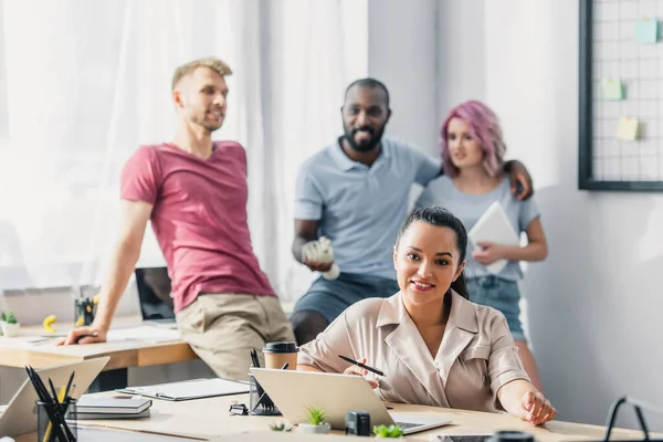 Focus selettivo della donna d'affari che guarda la fotocamera mentre lavora a tavola vicino ai colleghi multiculturali in ufficio — Foto stock