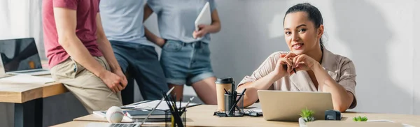 Panoramakonzept einer Geschäftsfrau, die den Stift in der Hand hält und in die Kamera schaut, während sie am Tisch in der Nähe von Kollegen im Büro arbeitet — Stockfoto