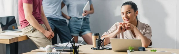 Panoramic concept of businesswoman holding pen and looking at camera while working at table near colleagues in office — Stock Photo