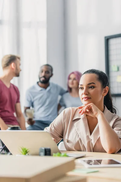 Selective focus of manager sitting near gadgets at table with multiethnic colleagues at background in office — Stock Photo