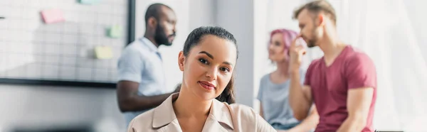 Imagen horizontal de la mujer de negocios mirando a la cámara con colegas multiculturales en el fondo de la oficina - foto de stock