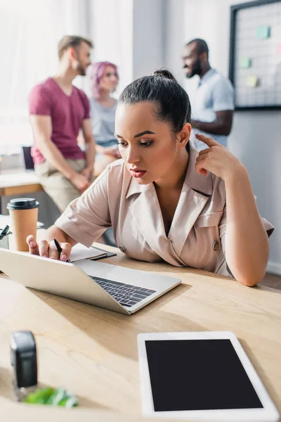 Selective focus of concentrated businesswoman using laptop with multicultural colleagues at background in office — Stock Photo