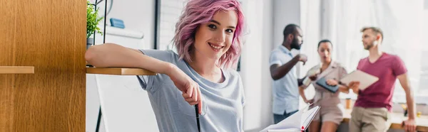 Panoramic shot of businesswoman holding pen near cupboard with multicultural colleagues at background in office — Stock Photo