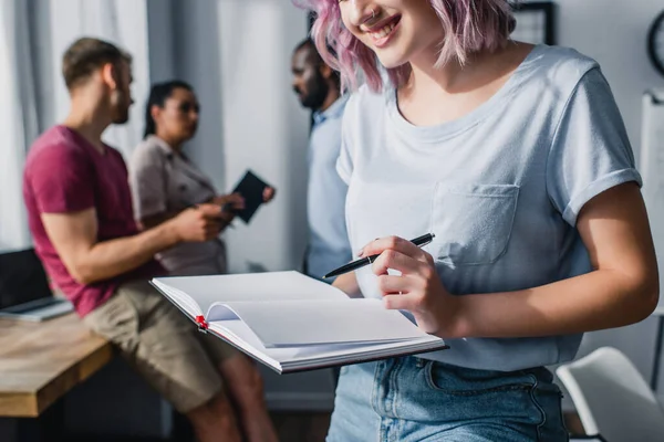 Selective focus of businesswoman holding notebook and pen while working near multiethnic colleagues in office — Stock Photo