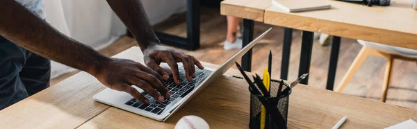 Orientación panorámica del hombre de negocios afroamericano utilizando portátil cerca de papelería y pelota de béisbol en la oficina - foto de stock