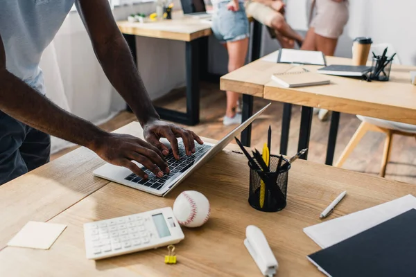 Vista recortada de un hombre de negocios americano fircan usando el ordenador portátil cerca de la calculadora y bola de béisbol en la mesa de la oficina - foto de stock