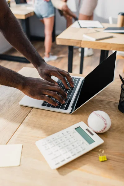 Cropped view of african american businessman using laptop with blank screen near calculator and baseball ball on office table — Stock Photo