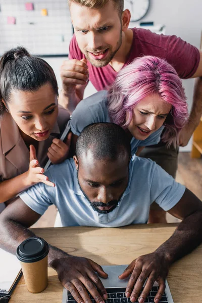 Empresários multiculturais concentrados olhando para colega afro-americano usando laptop à mesa no escritório — Stock Photo