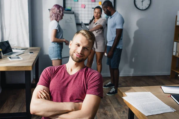 Selective focus of businessman with crossed arms looking at camera while multiethnic colleagues working at background — Stock Photo