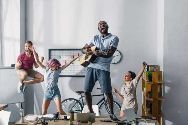 Селективный фокус африканского американского бизнесмена, играющего на акустической гитаре, в то время как мультиэтнические коллеги танцуют в офисе — стоковое фото