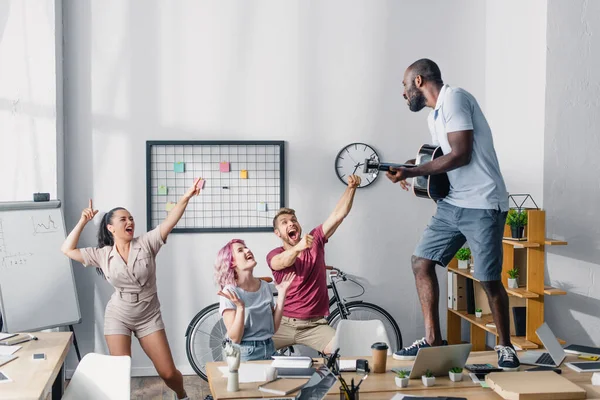 Selektiver Fokus eines afrikanisch-amerikanischen Geschäftsmannes, der Akustikgitarre spielt, während multikulturelle Mitarbeiter im Büro tanzen — Stockfoto