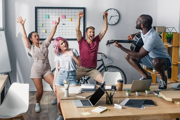 Selective focus of business people dancing near african american colleague with acoustic guitar in office — Stock Photo