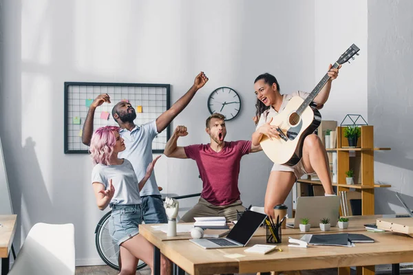 Selective focus of businesswoman playing acoustic guitar near multicultural colleagues singing and dancing in office — Stock Photo