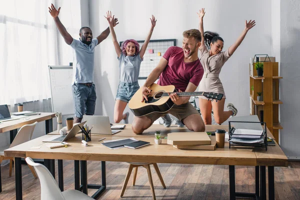 Selective focus of multiethnic business people jumping while colleague playing acoustic guitar in office — Stock Photo