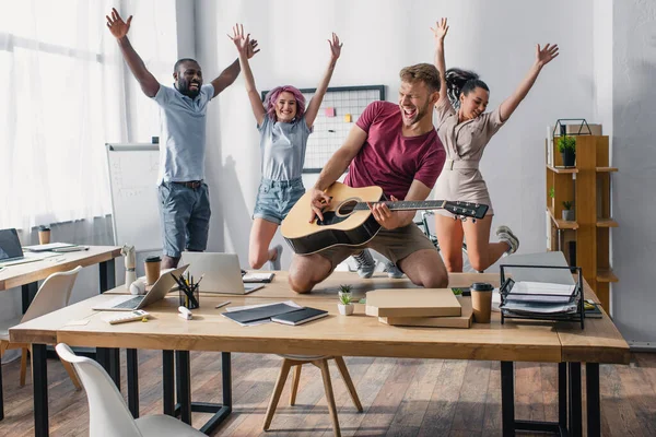 Enfoque selectivo de gente de negocios multiétnicos saltando mientras su colega toca la guitarra acústica en la oficina - foto de stock