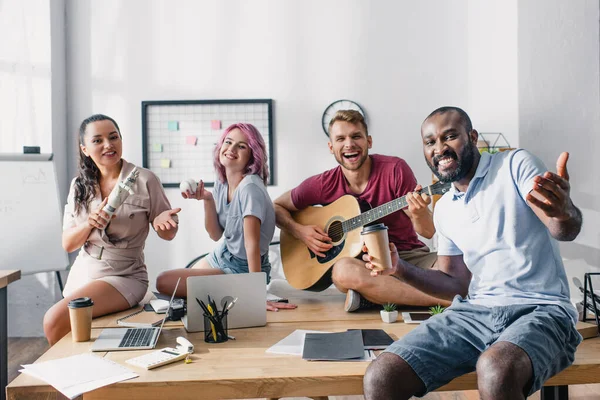 Jovens empresários multiétnicos com bola de beisebol, café para ir e guitarra acústica olhando para a câmera no escritório — Fotografia de Stock