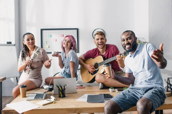 Jeunes hommes d'affaires multiethniques avec balle de baseball, café à emporter et guitare acoustique regardant la caméra au bureau — Photo de stock