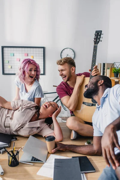 Вибірковий фокус сміху багатокультурних бізнесменів з акустичною гітарою, сидячи на столі в офісі — стокове фото