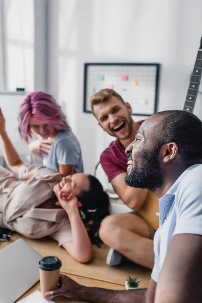 Selective focus of african american businessman holding coffee to go near multiethnic colleagues with acoustic guitar on table — Stock Photo