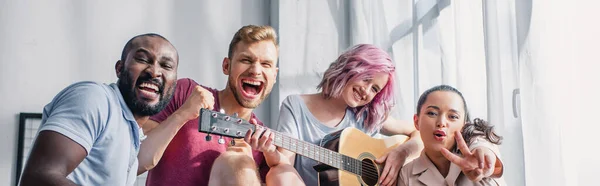 Prise de vue panoramique de jeunes hommes d'affaires multiethniques avec guitare acoustique regardant la caméra au bureau — Photo de stock