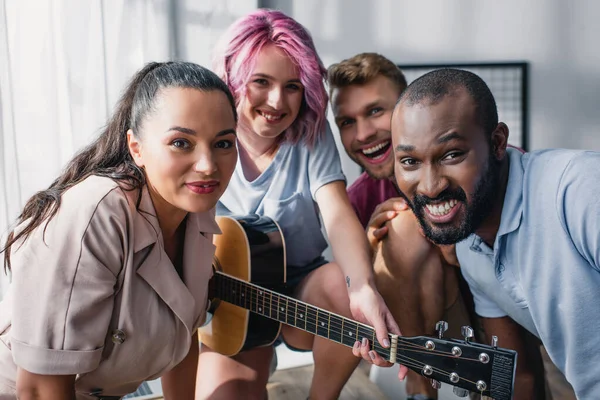 Enfoque selectivo de personas de negocios multiculturales con guitarra acústica mirando a la cámara en la oficina - foto de stock